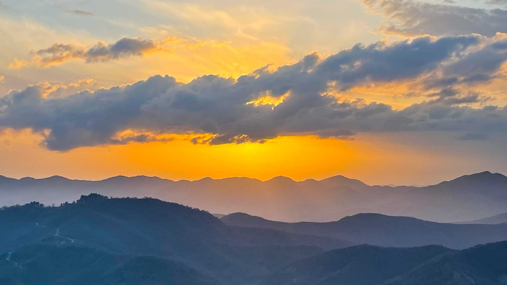 A vibrant sunset illuminates the Albanian mountains with hues of orange and yellow, under a partly cloudy sky.