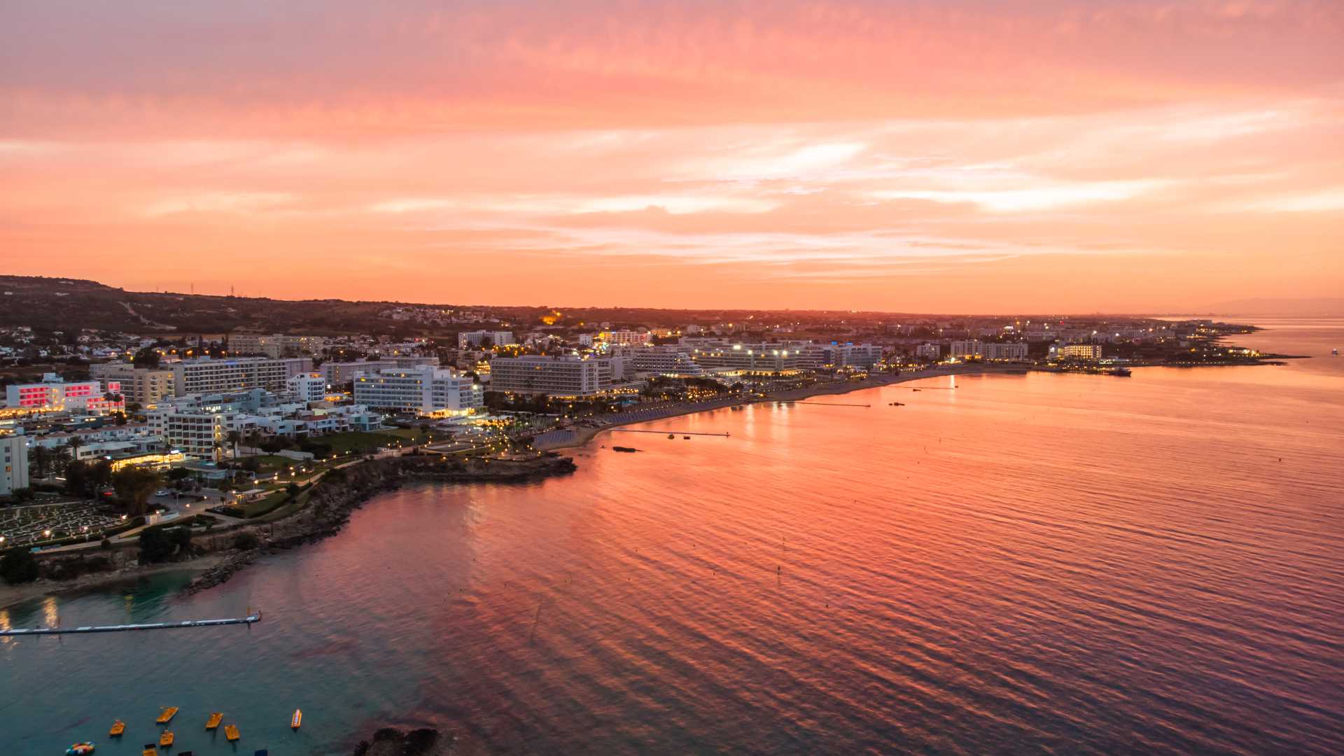 Sonnenuntergang über Protaras, Zypern, mit den Lichtern der Stadt, die sich im ruhigen Mittelmeerwasser spiegeln.