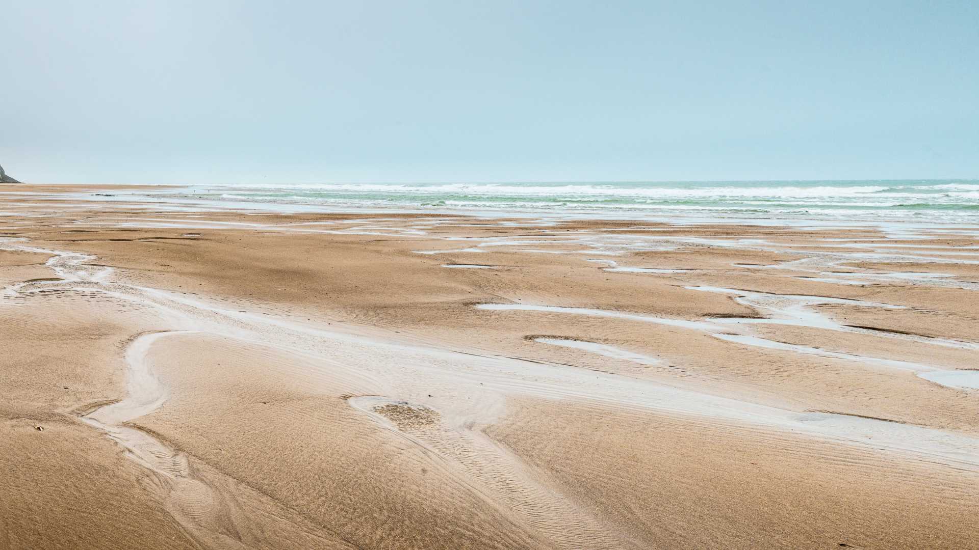 Weitläufiger Sandstrand von Cap Blanc-Nez mit sanften Wellen und einer entfernten Klippe unter einem klaren, hellblauen Himmel.