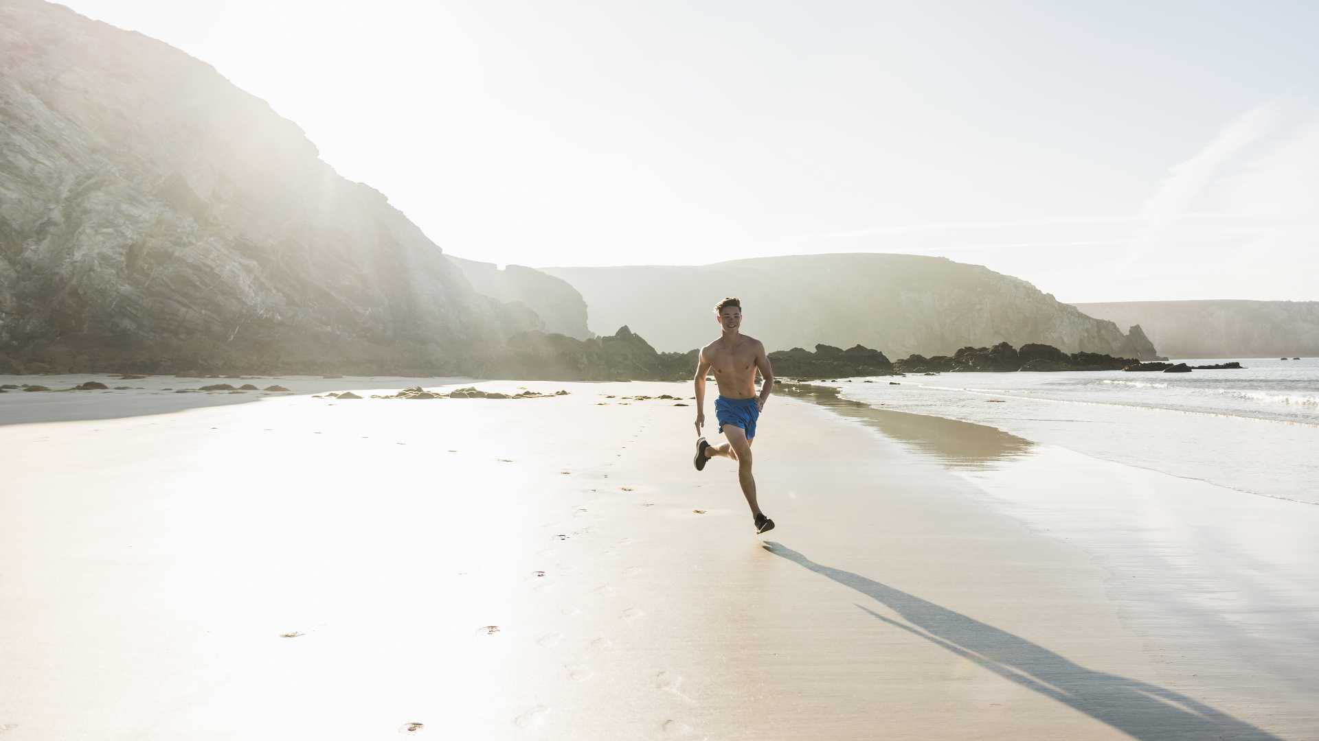 Een jonge man rent shirtloos over een sereen strand van het schiereiland Crozon, badend in zacht zonlicht.