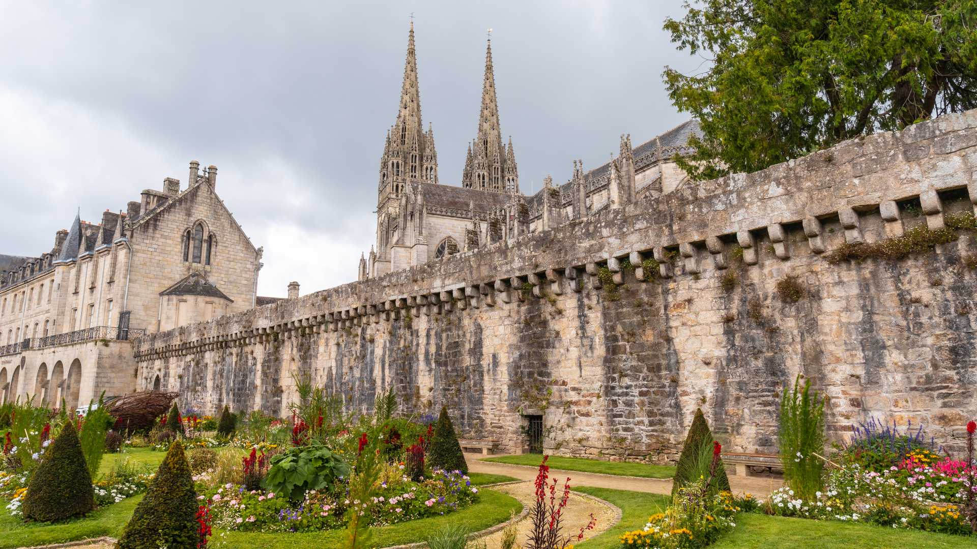 Middeleeuwse muren en de torenspitsen van de Saint Corentin kathedraal in Quimper, omringd door levendige tuinen.