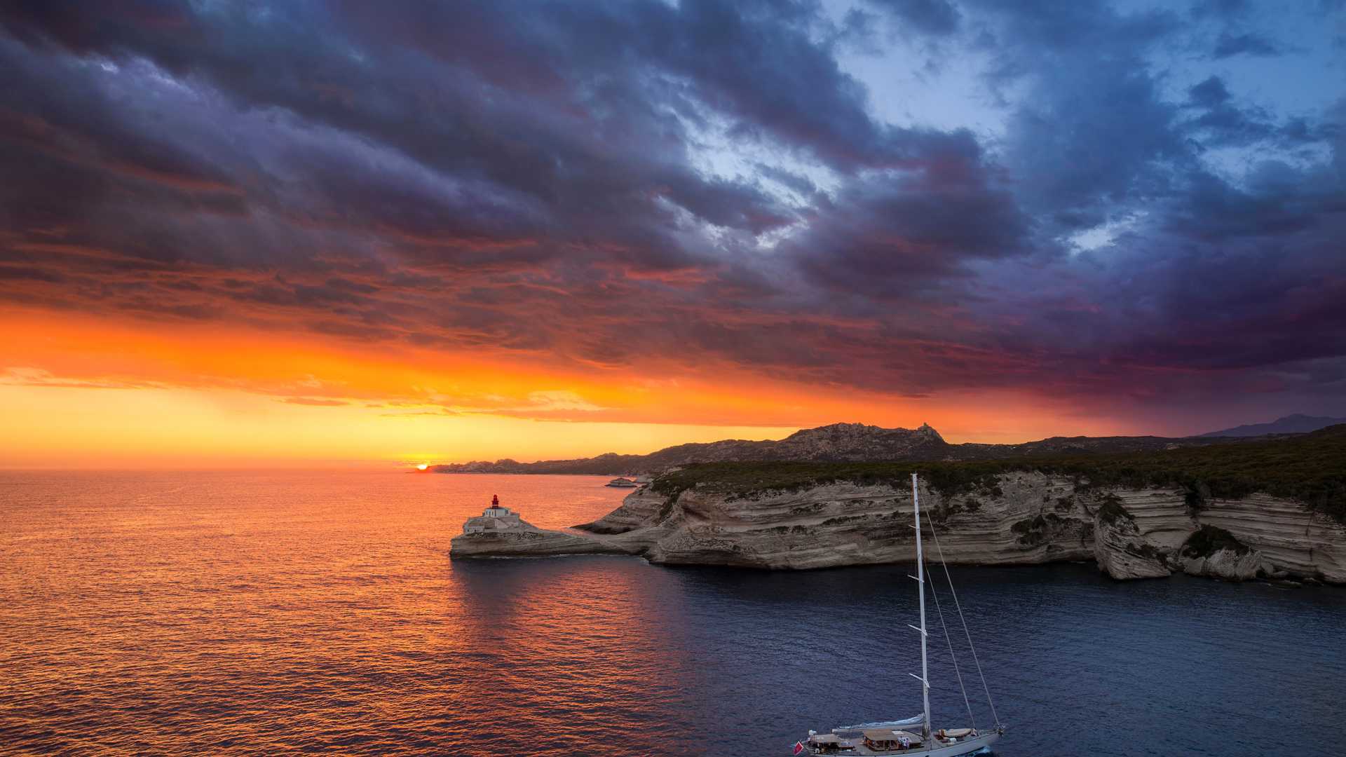 Fiery sunset over Bonifacio lighthouse, with dramatic clouds and serene sea, Corsica, France.