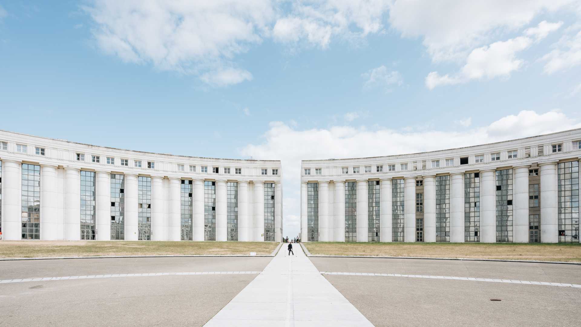 Geschwungene weiße Gebäude mit hohen Säulen unter blauem Himmel bei Axe Majeur in Cergy, Frankreich.