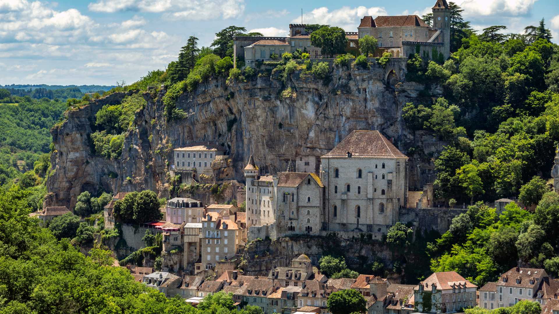 In Rocamadour fallen mittelalterliche Gebäude kaskadenförmig von einer Klippe herab, umgeben von üppigem Grün und einem strahlenden Himmel.