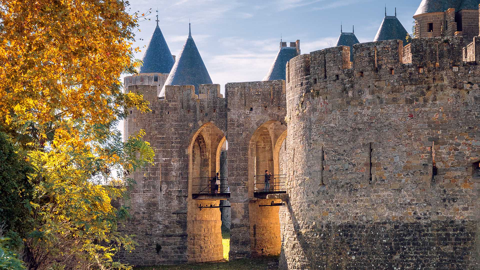 Entrada medieval de Porte Narbonnaise a Carcassonne, enmarcada por árboles de otoño y antiguas murallas de piedra.