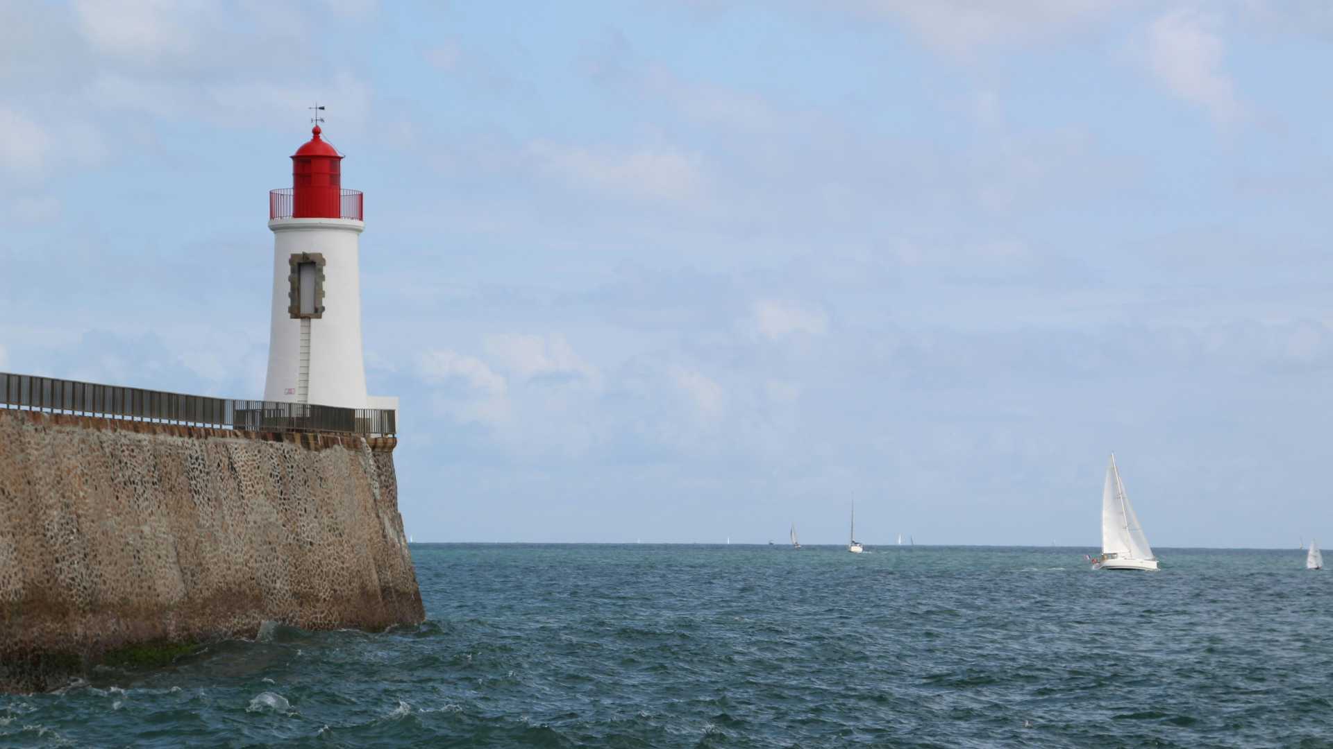 Phare blanc avec un sommet rouge aux Sables-d'Olonne, France, surplombant une mer sereine avec des voiliers.