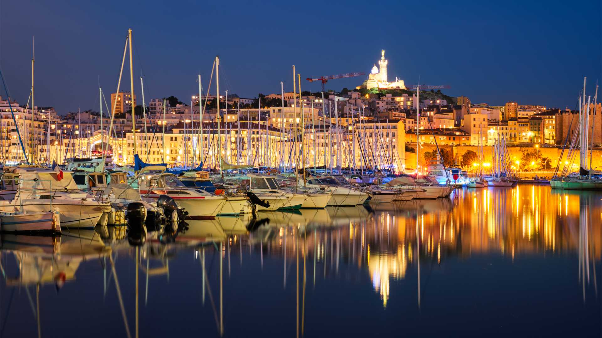 Yachts are mirrored in the tranquil waters of Marseille's Old Port at night, with the illuminated Basilica of Notre-Dame de la Garde in the background.