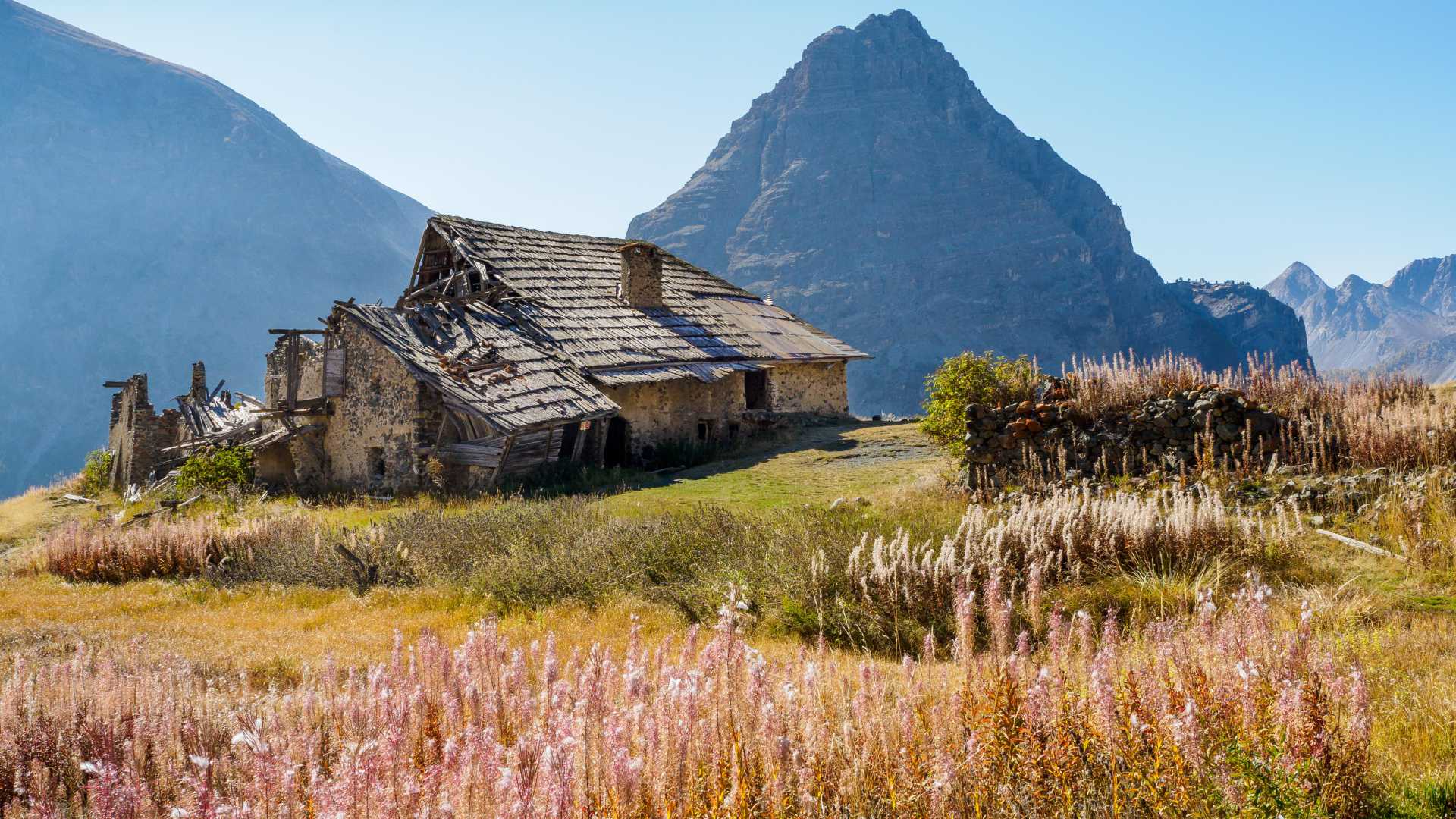 Una casa de piedra abandonada en Les Fraches, rodeada de flores silvestres con imponentes picos alpinos al fondo.