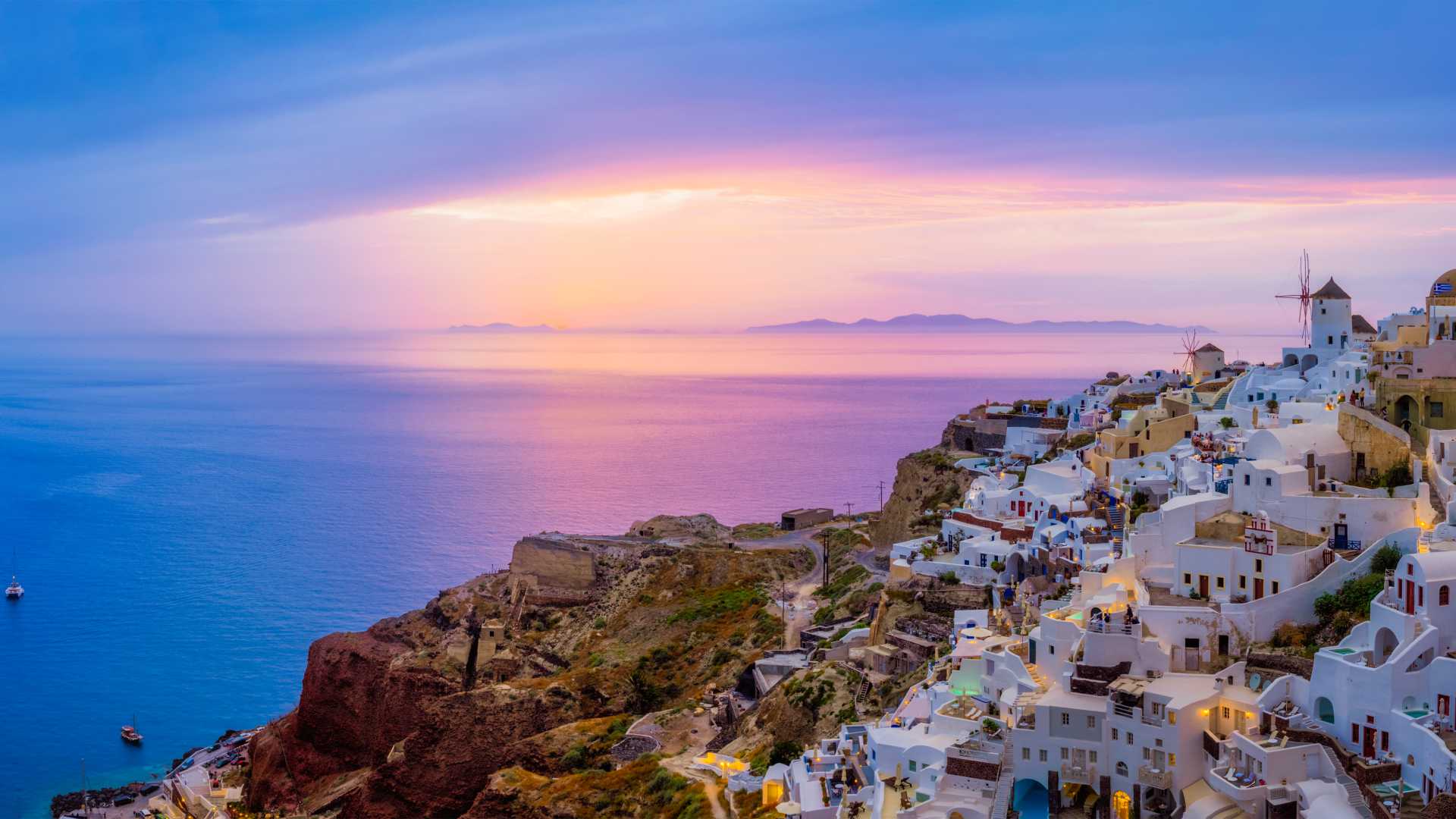 Oia village with white houses and windmills overlooks a serene sunset over the Aegean Sea in Santorini, Greece.