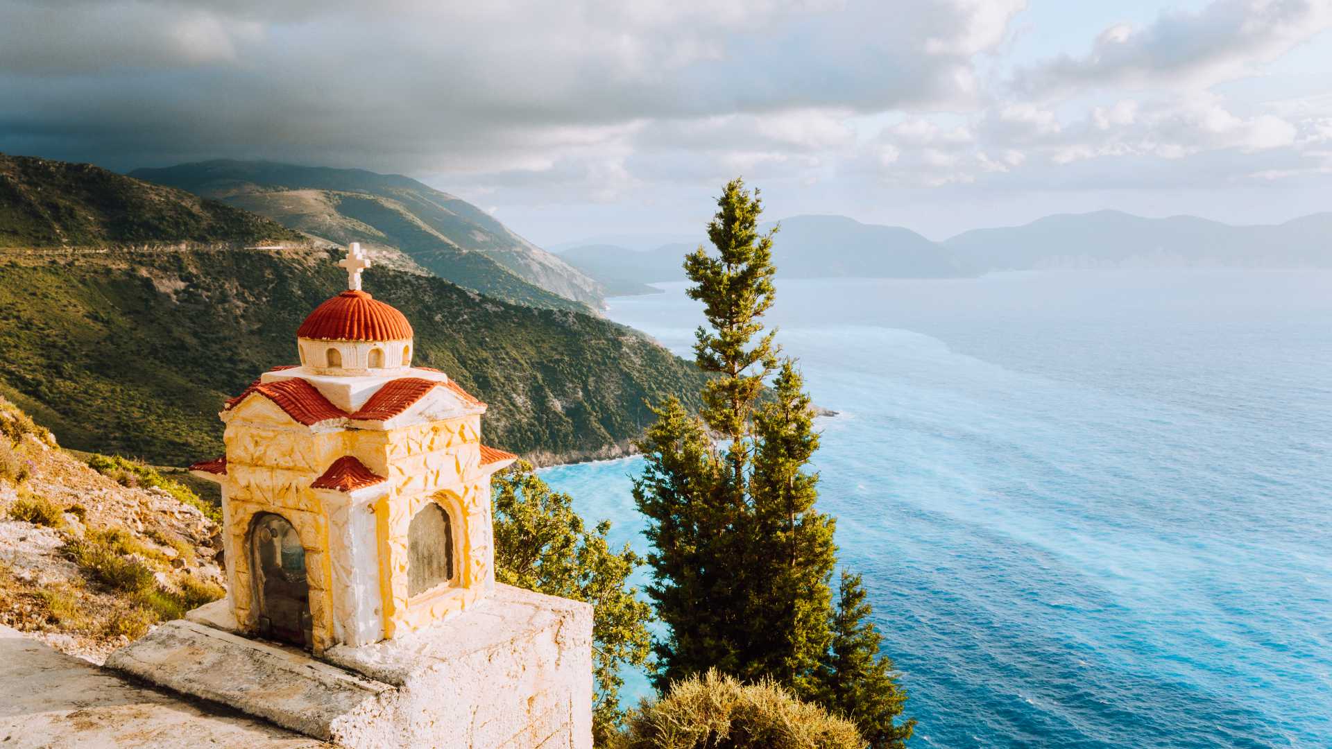 Colorful Proskinitari shrine on a cliff's edge with a stunning coastline and cloudscape in the background.