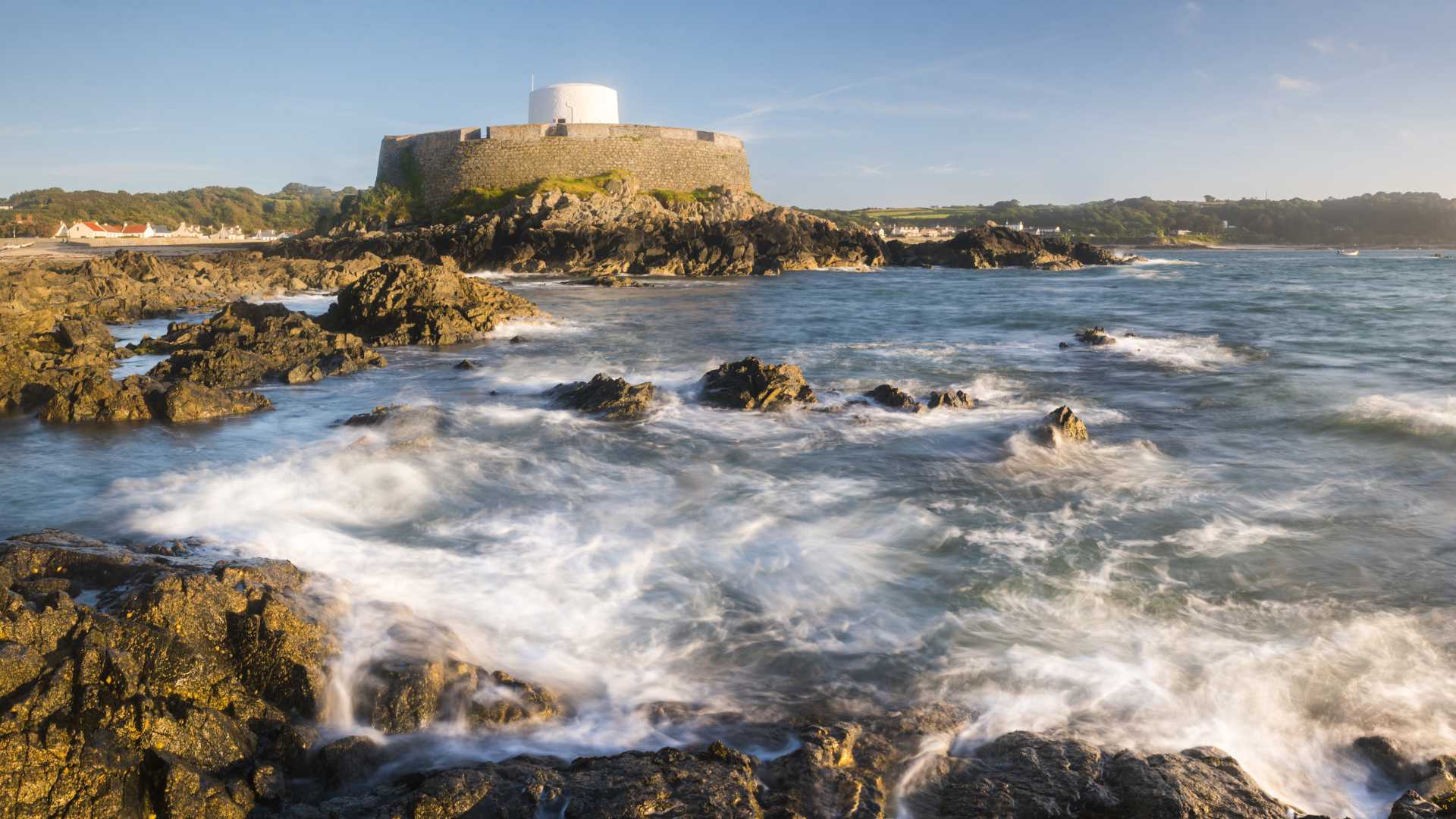 Fort Grey aan de rotsachtige kust van Guernsey bij zonsondergang, met golven die tegen de ruige kustlijn slaan.