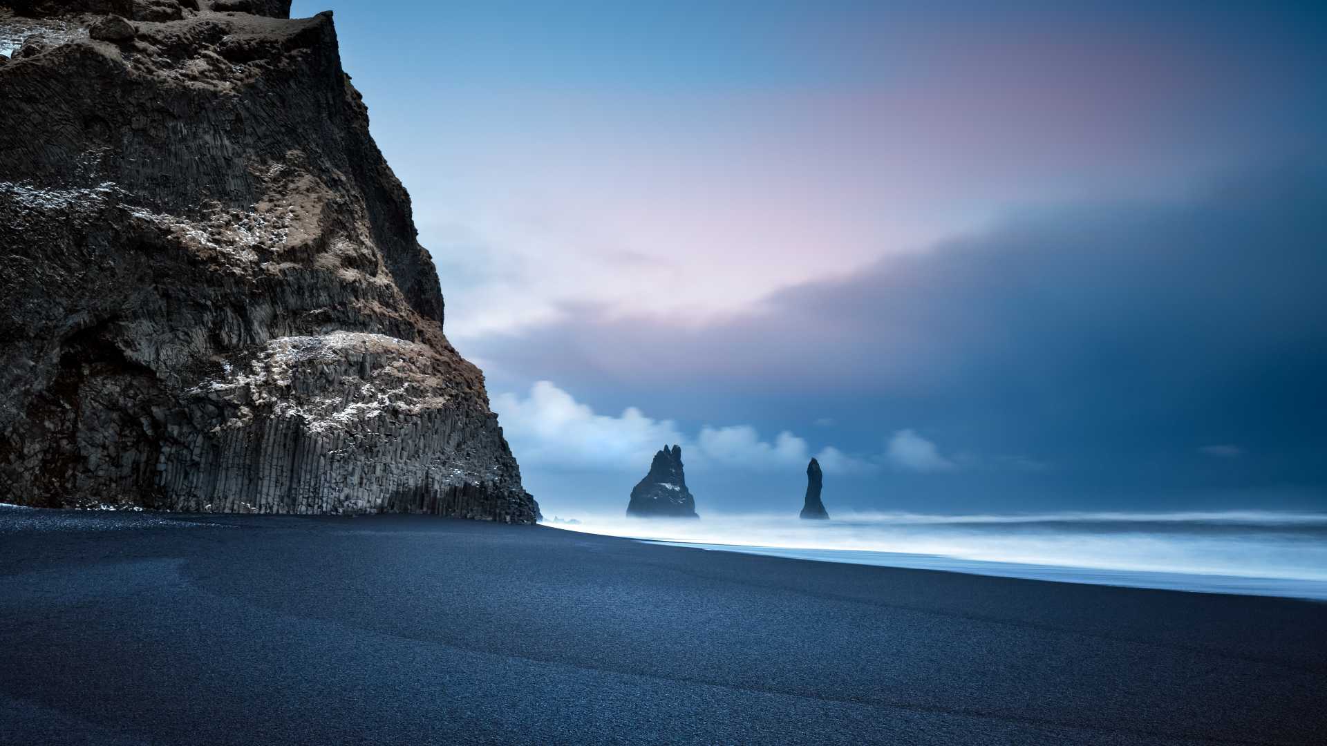 Des falaises de basalte et des falaises maritimes s'élèvent au-dessus de la plage de sable noir de Vik, en Islande, sous un ciel pastel.