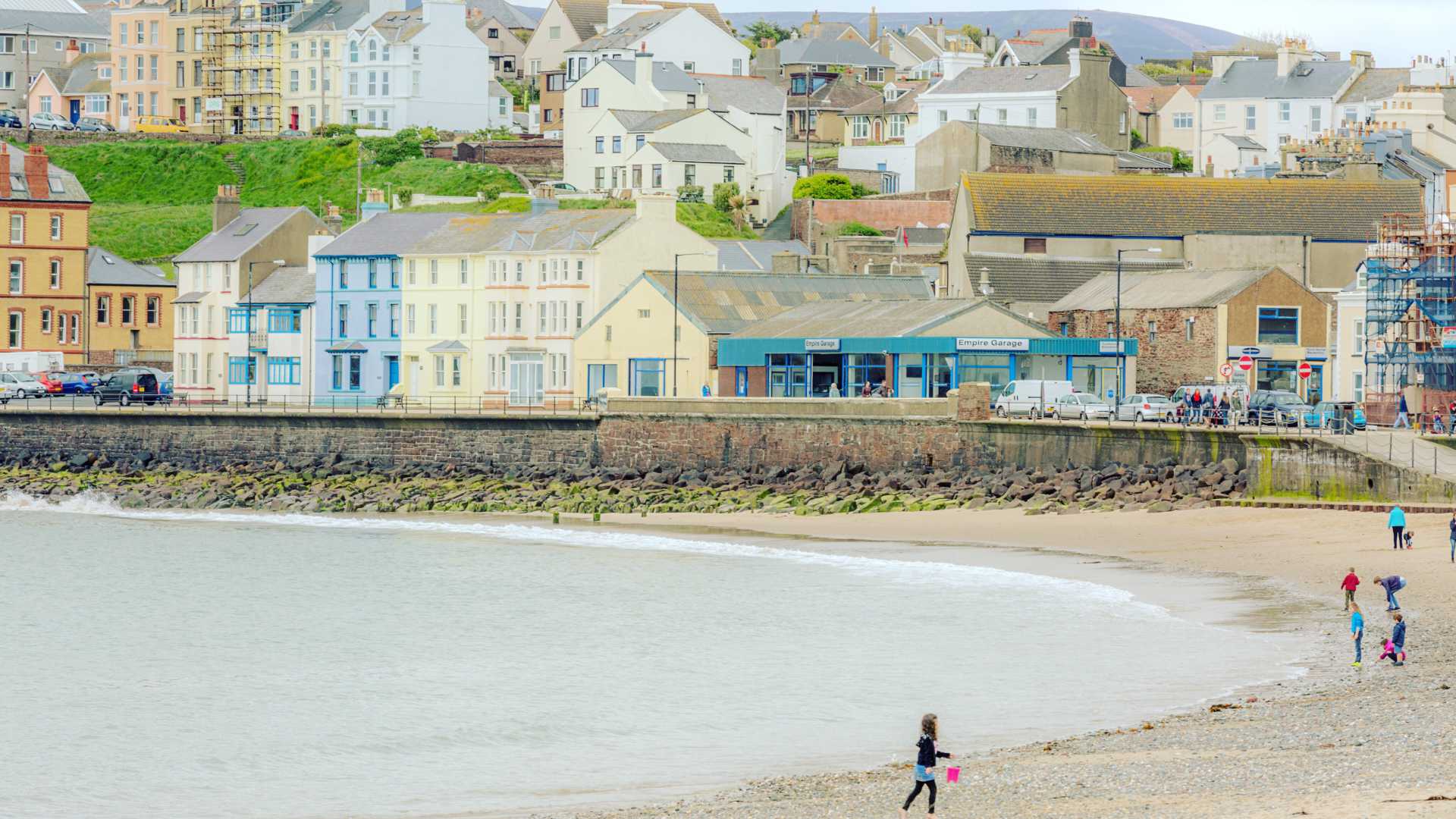 Colorful buildings line the shoreline of a sandy beach in Peel, Isle of Man, with people strolling along.