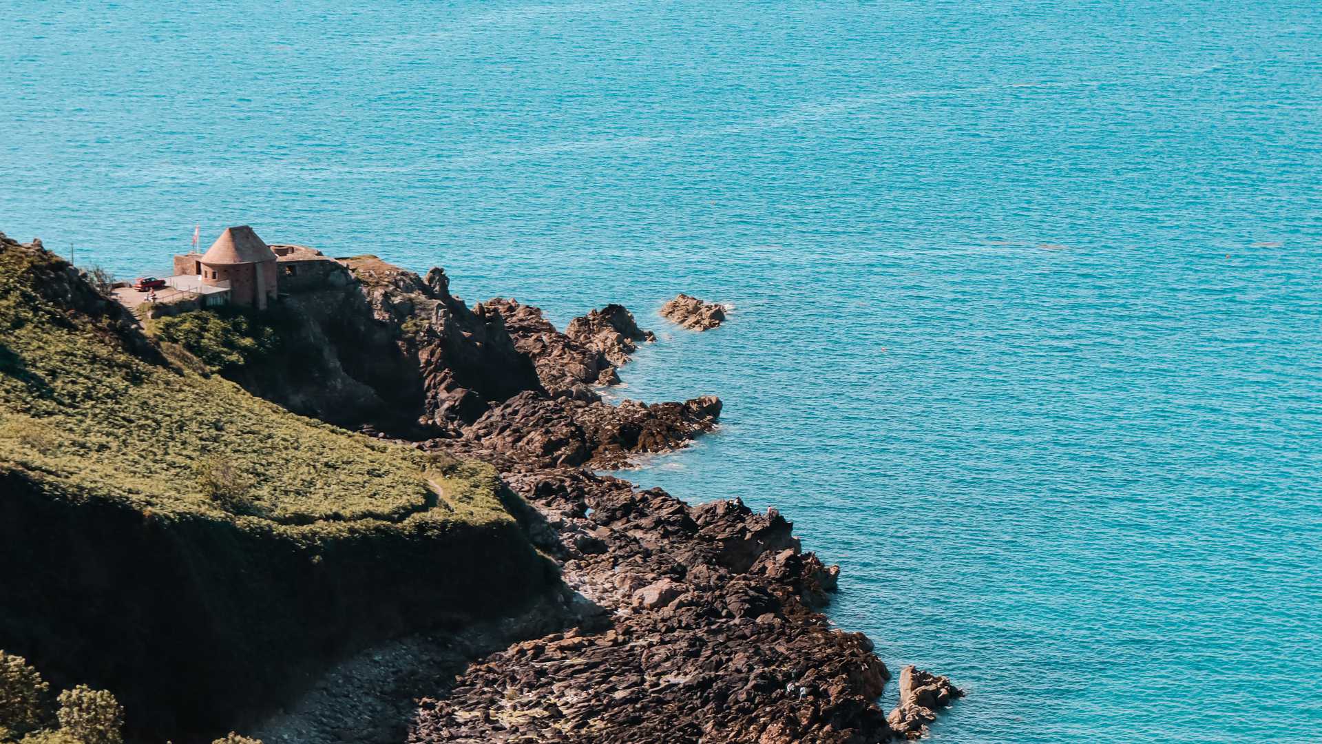 A rugged coastline with a small stone building overlooking the blue waters of the sea on Jersey shore.