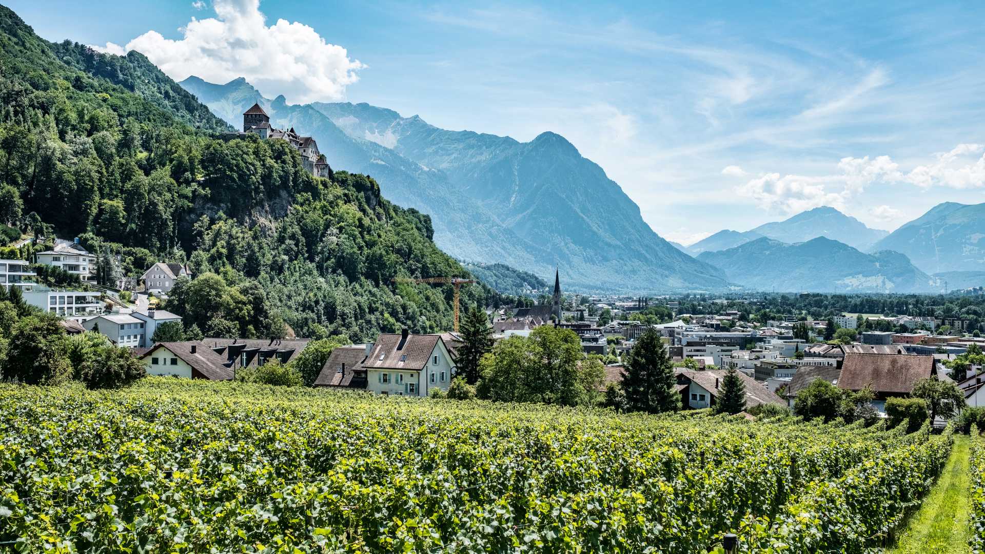 A scenic view of Vaduz with a hilltop castle, lush greenery, and the majestic Alps in the background.