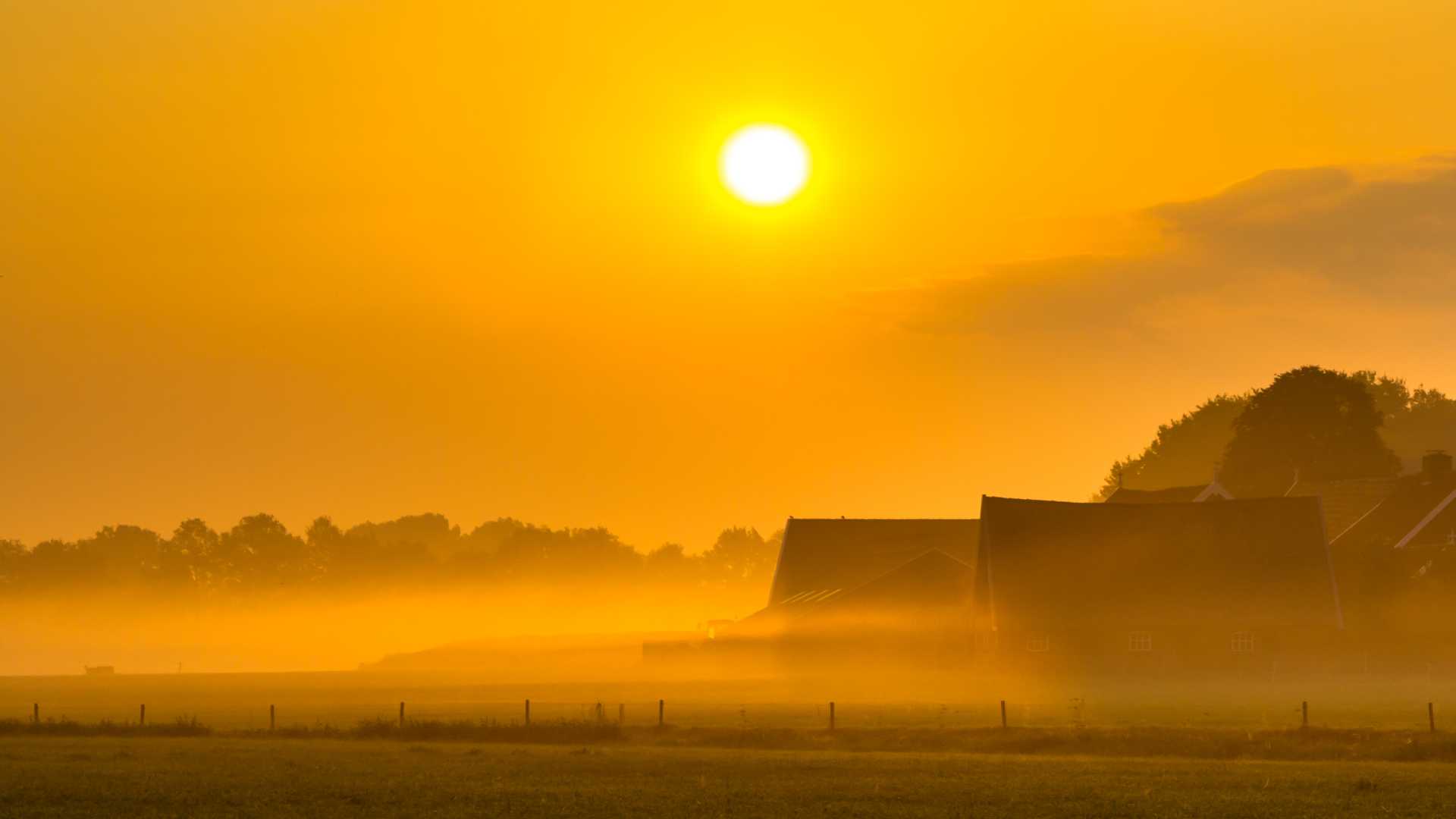 Amanecer iluminando un brumoso paisaje agrícola con graneros y un tractor