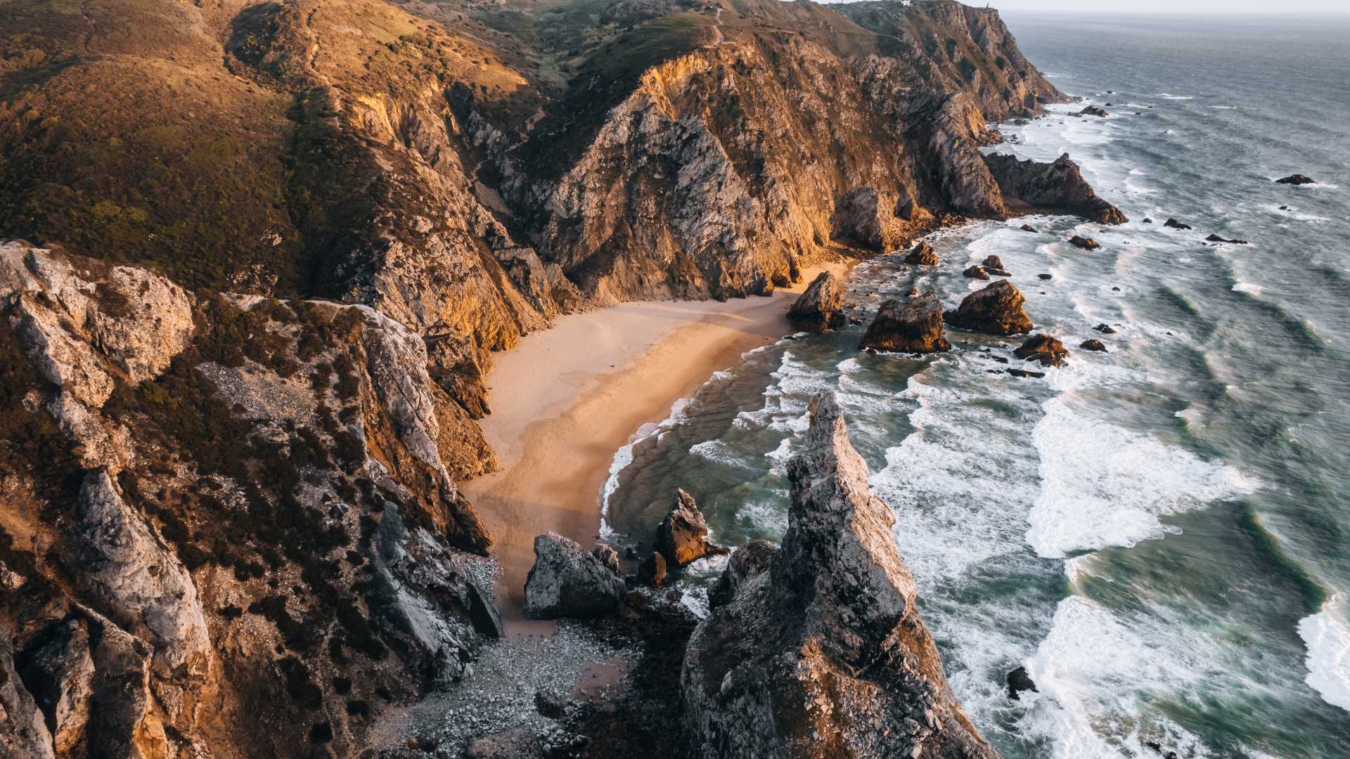 Aerial view of Praia da Ursa's dramatic cliffs and secluded beach at sunset, waves crashing along the rugged coast.