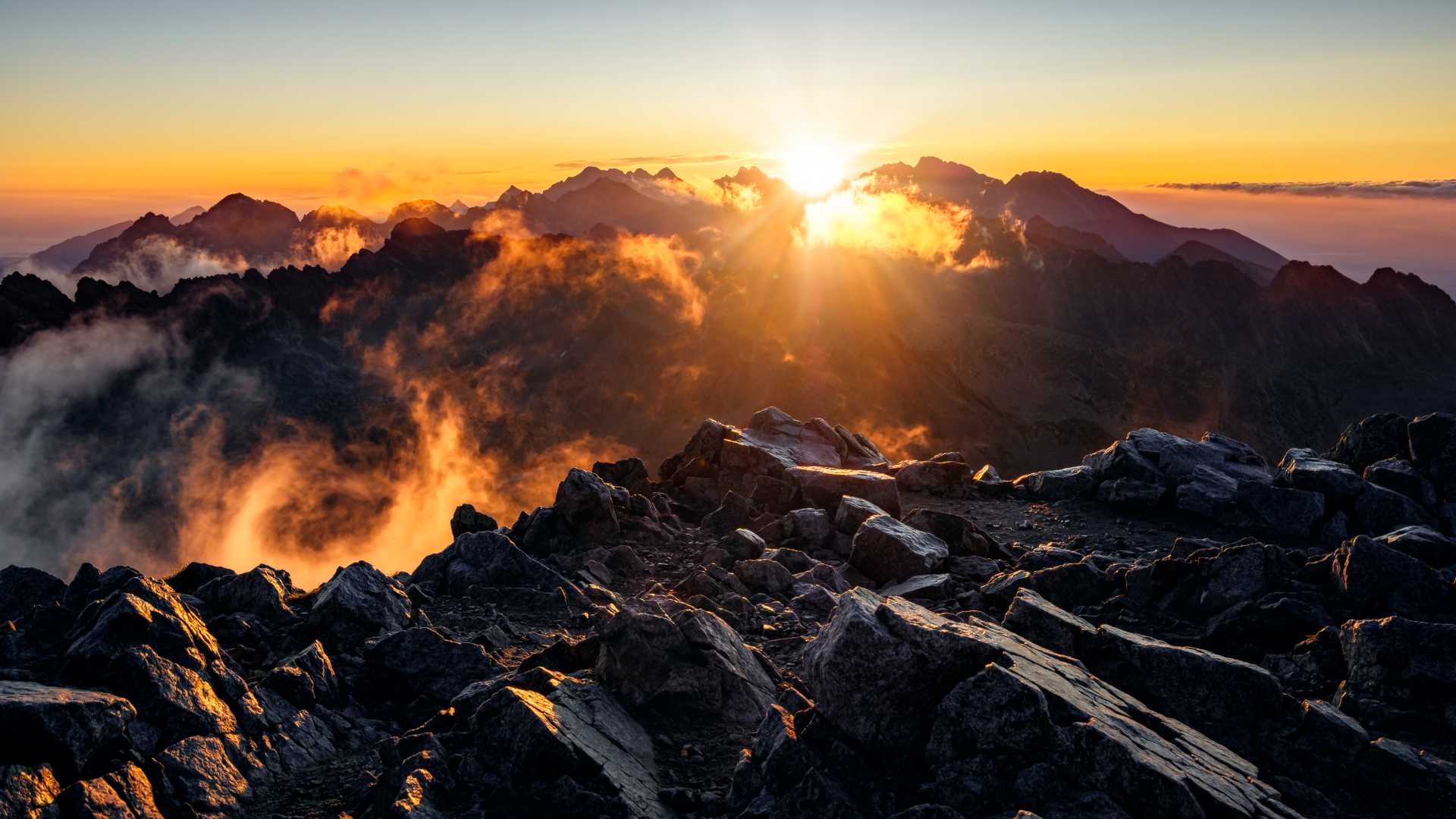 Ein goldener Sonnenaufgang beleuchtet die felsigen Gipfel der Hohen Tatra in der Slowakei, wobei die Wolken einen dramatischen Akzent setzen.