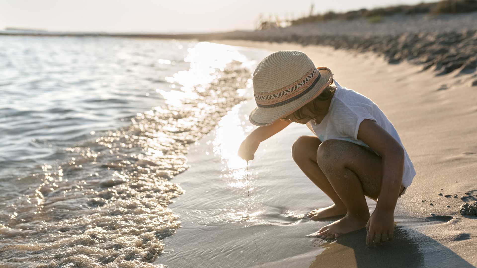 Een klein meisje met een zonnehoed speelt bij de glinsterende kust van een Menorca strand bij zonsondergang, Spanje.