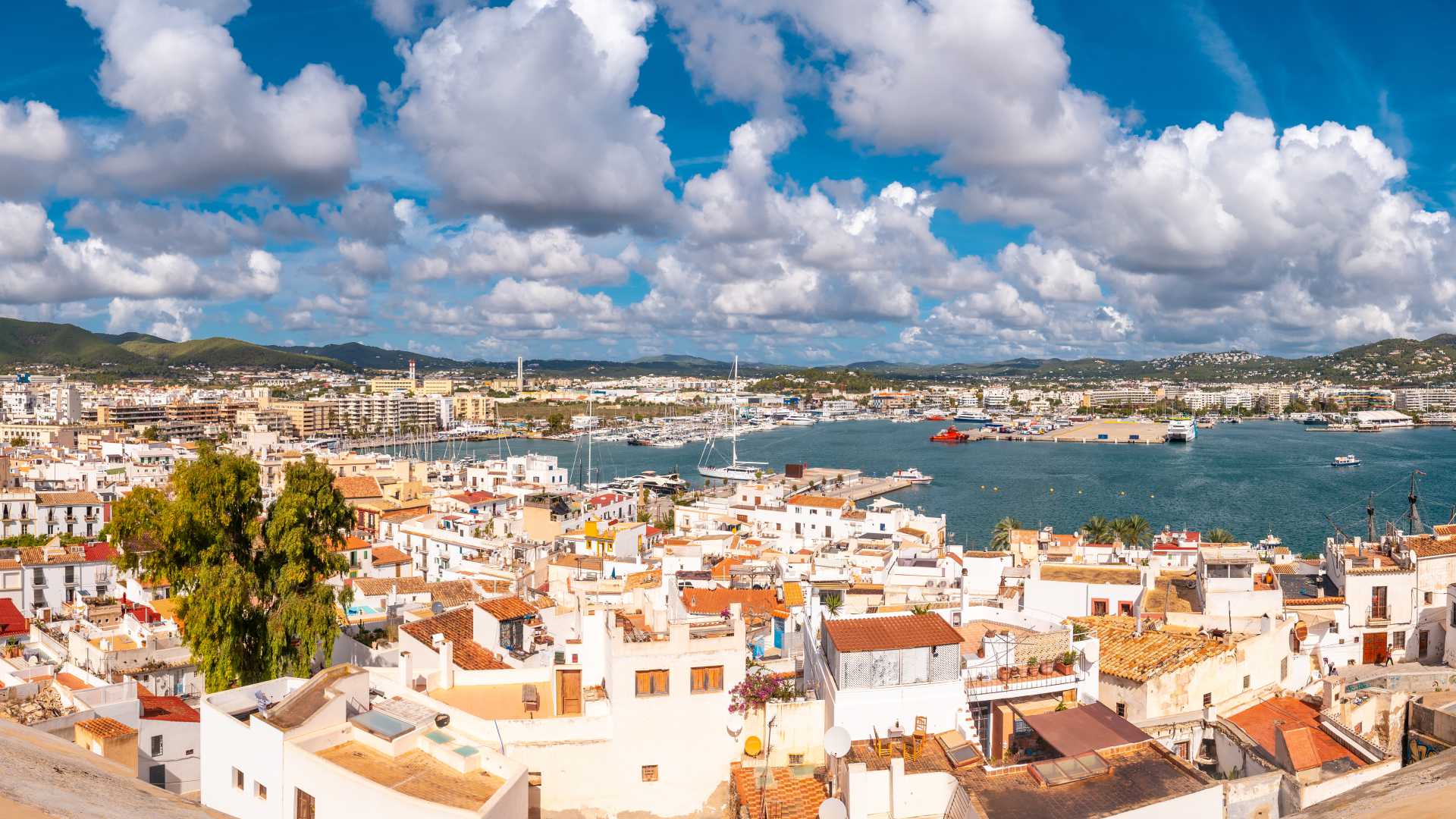 A panoramic view of Ibiza's old town and bustling port, framed by a vibrant blue sky with fluffy clouds.