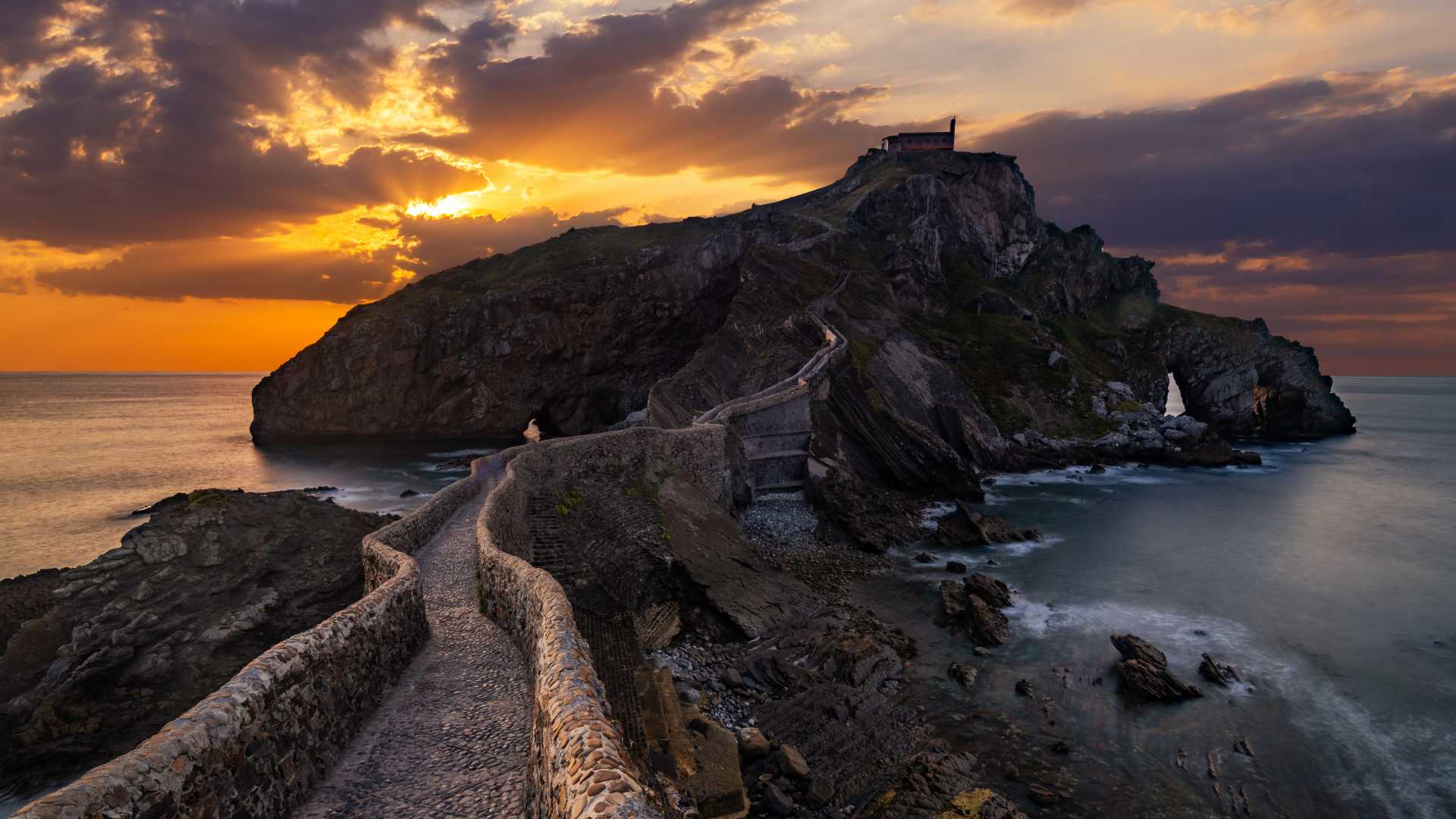 Een kronkelend stenen pad leidt naar San Juan de Gaztelugatxe, met een dramatische zonsondergang over het rotsachtige eiland.