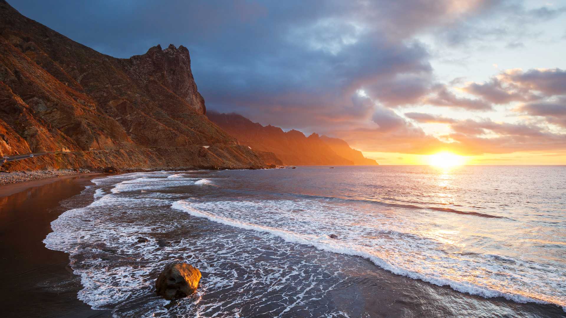 Coucher de soleil doré sur la plage de sable noir de Tenerife, avec ses falaises abruptes et ses vagues qui s'écrasent sur le rivage.