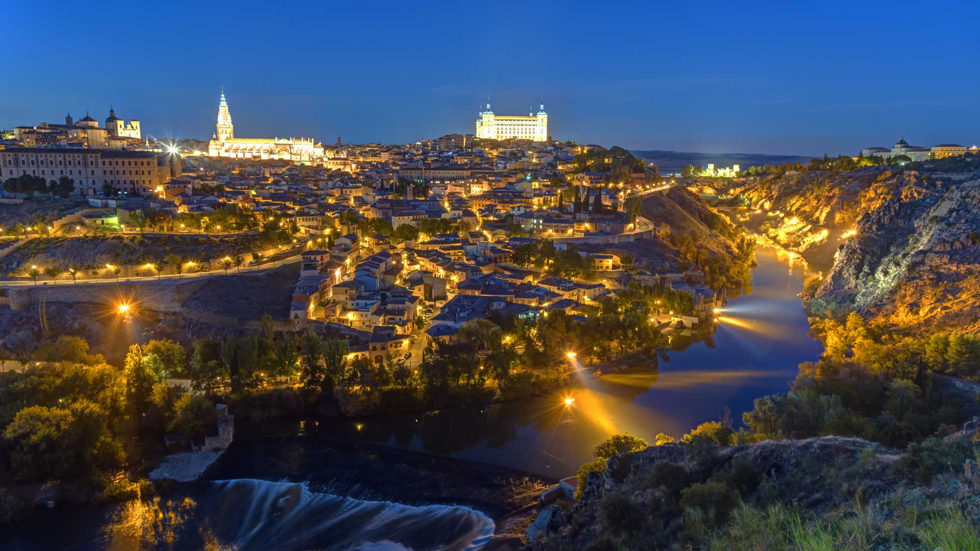 De historische oude stad van Toledo gloeit 's nachts, met het Alcázar en de kathedraal verlicht. De rivier de Taag kronkelt eronder.