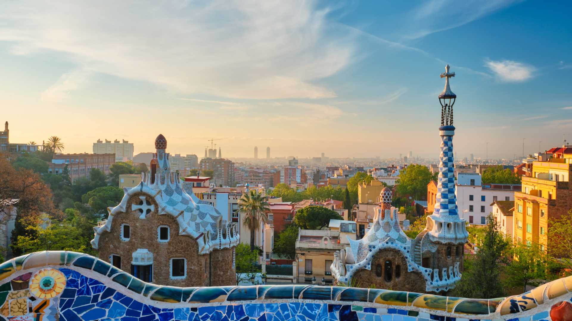 Colorful mosaic buildings in Park Guell with Barcelona skyline at sunrise.