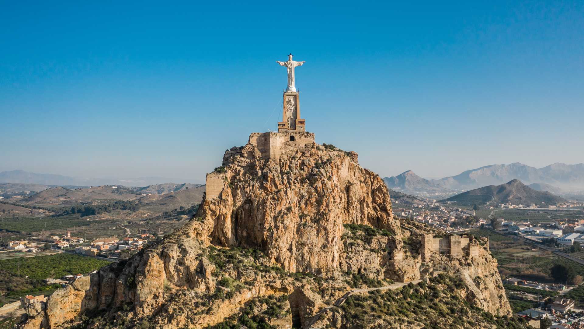 Luchtfoto van het kasteel van Monteagudo, bekroond door een Christusbeeld, met het ruige landschap van Murcia op de achtergrond.