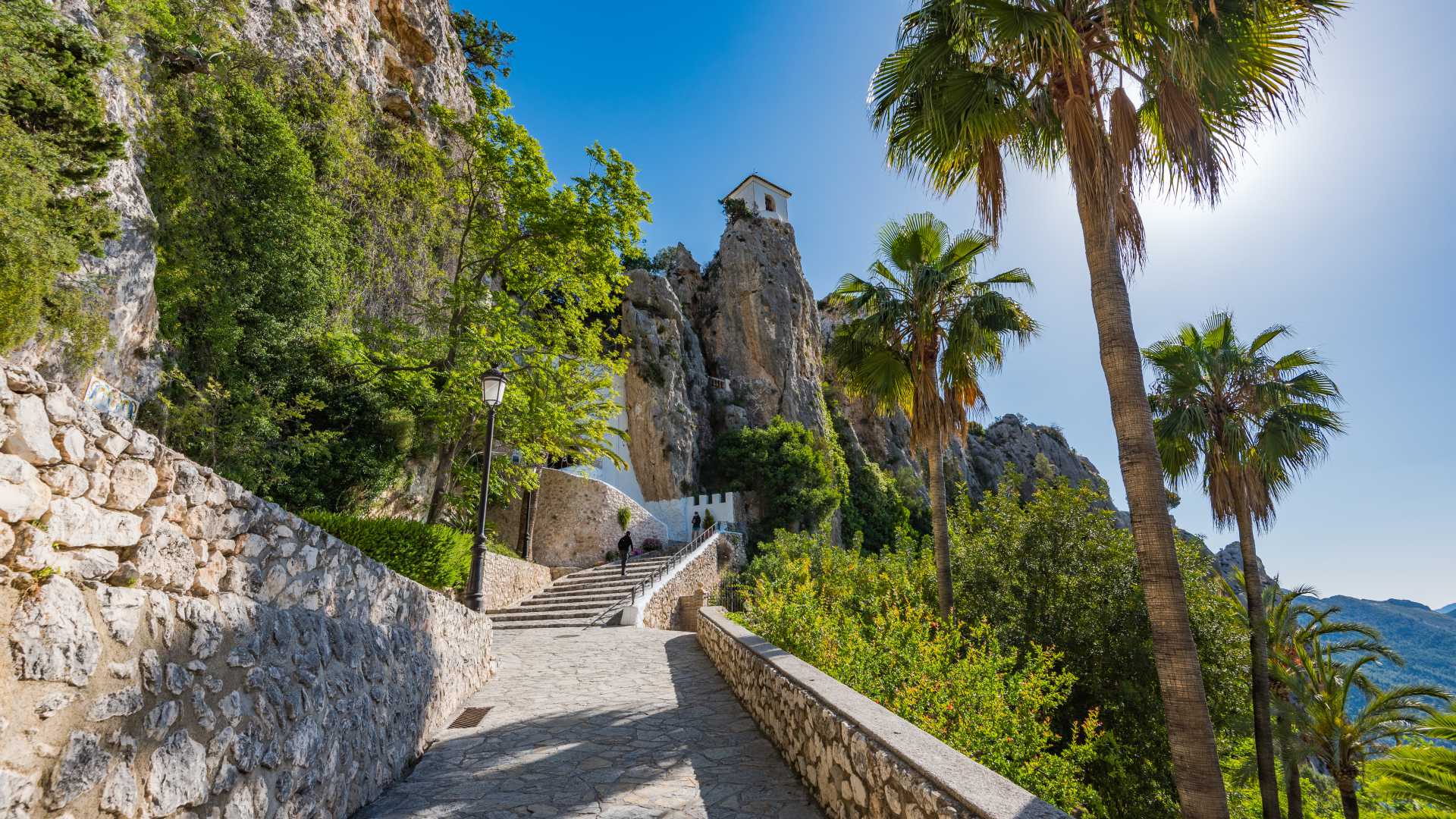 A stone path leads to Guadalest Castle perched on a rocky cliff, surrounded by lush greenery and palm trees.