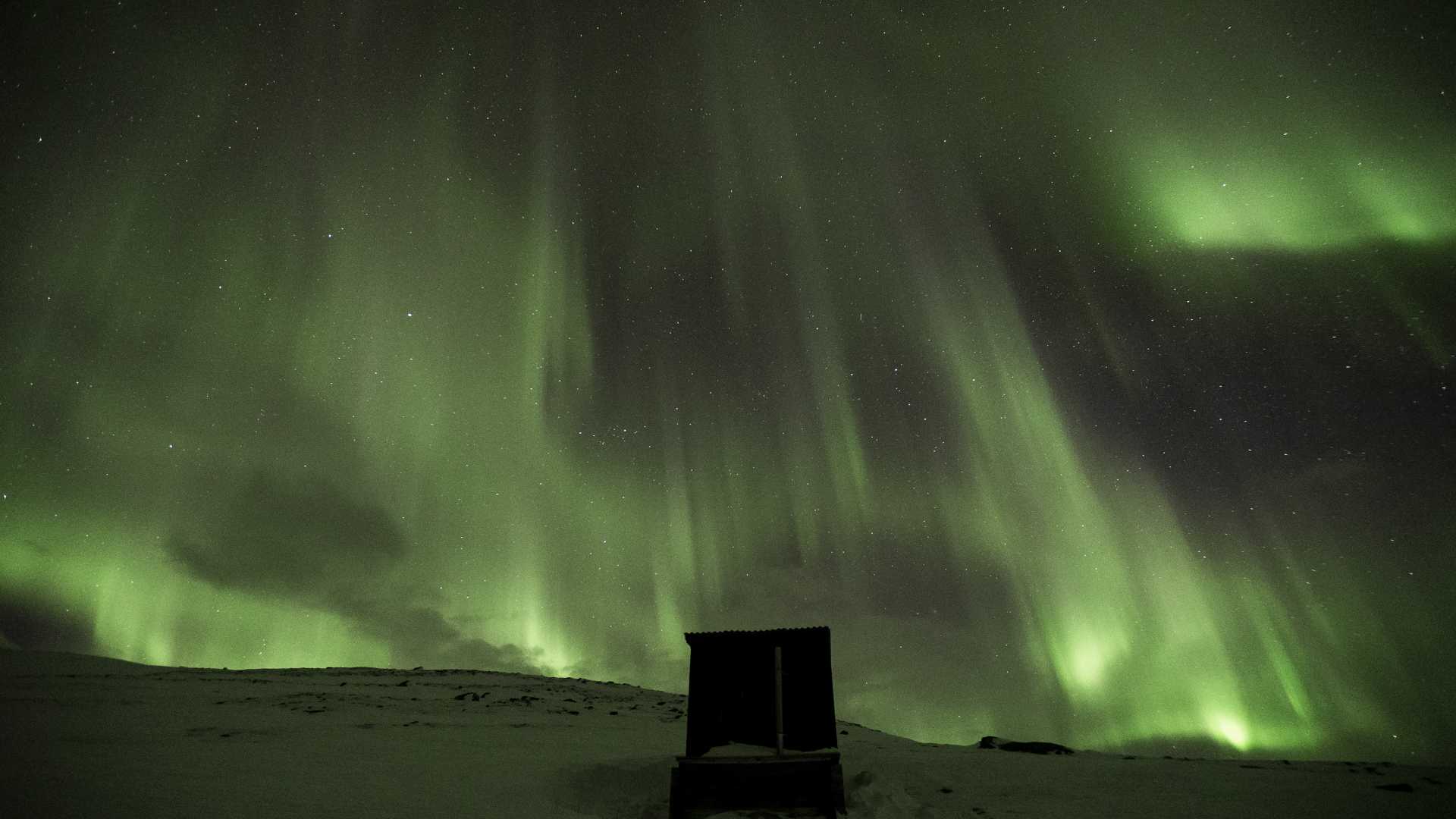 Northern lights illuminate the night sky over a snowy landscape in Abisko, Sweden, with a small hut below.