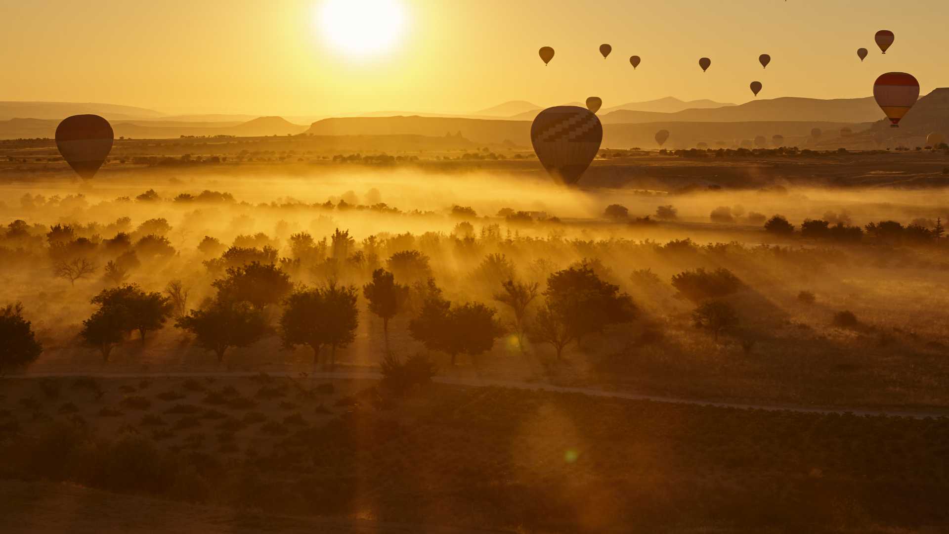 Heißluftballons schweben in der Abenddämmerung über die goldene Landschaft Kappadokiens und werfen Schatten auf das neblige Tal darunter.