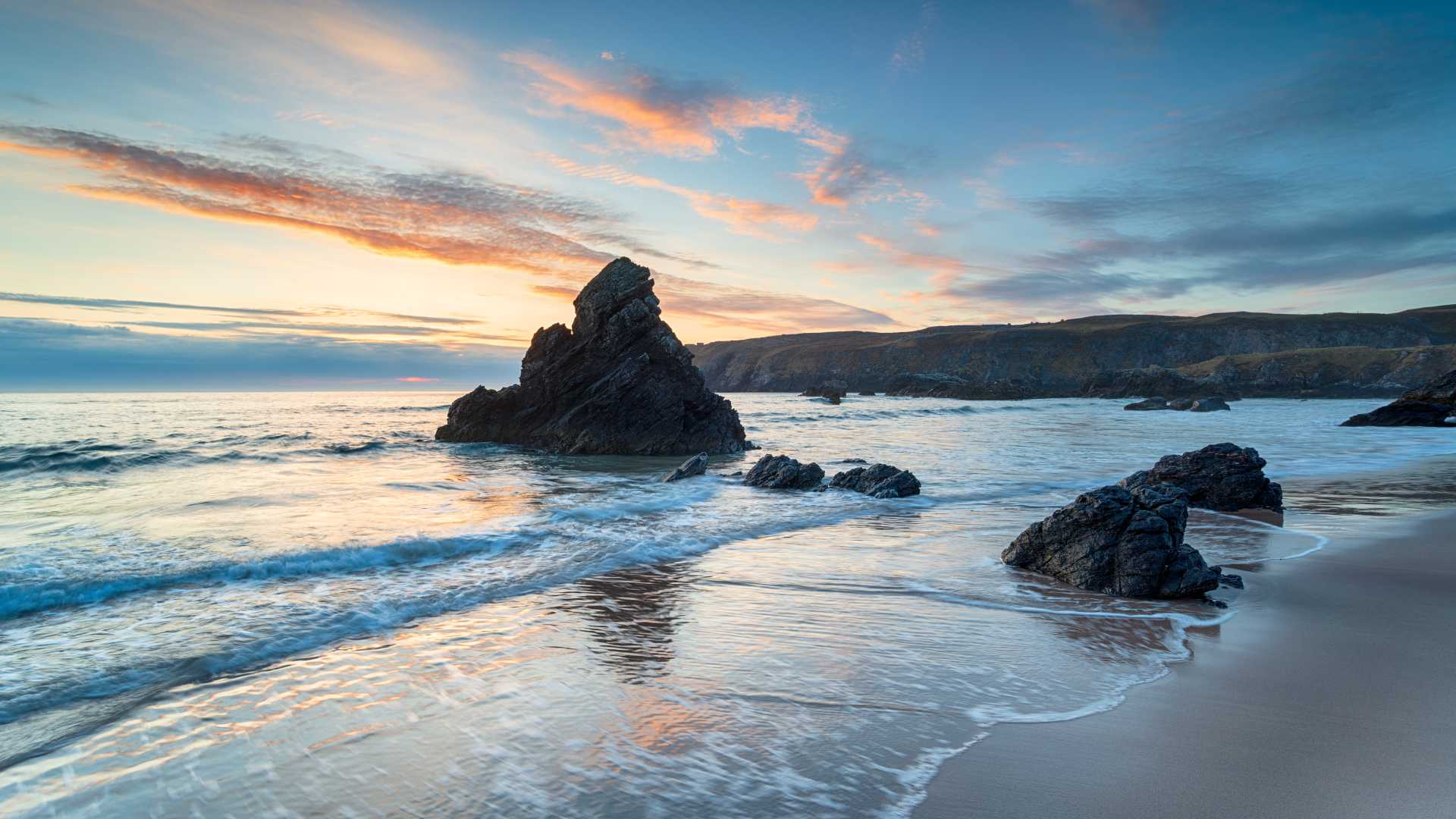 Zonsopgang boven Sango Bay in Durness, Schotland, met zeestapels en zachte golven die tegen de zandkust kabbelen.