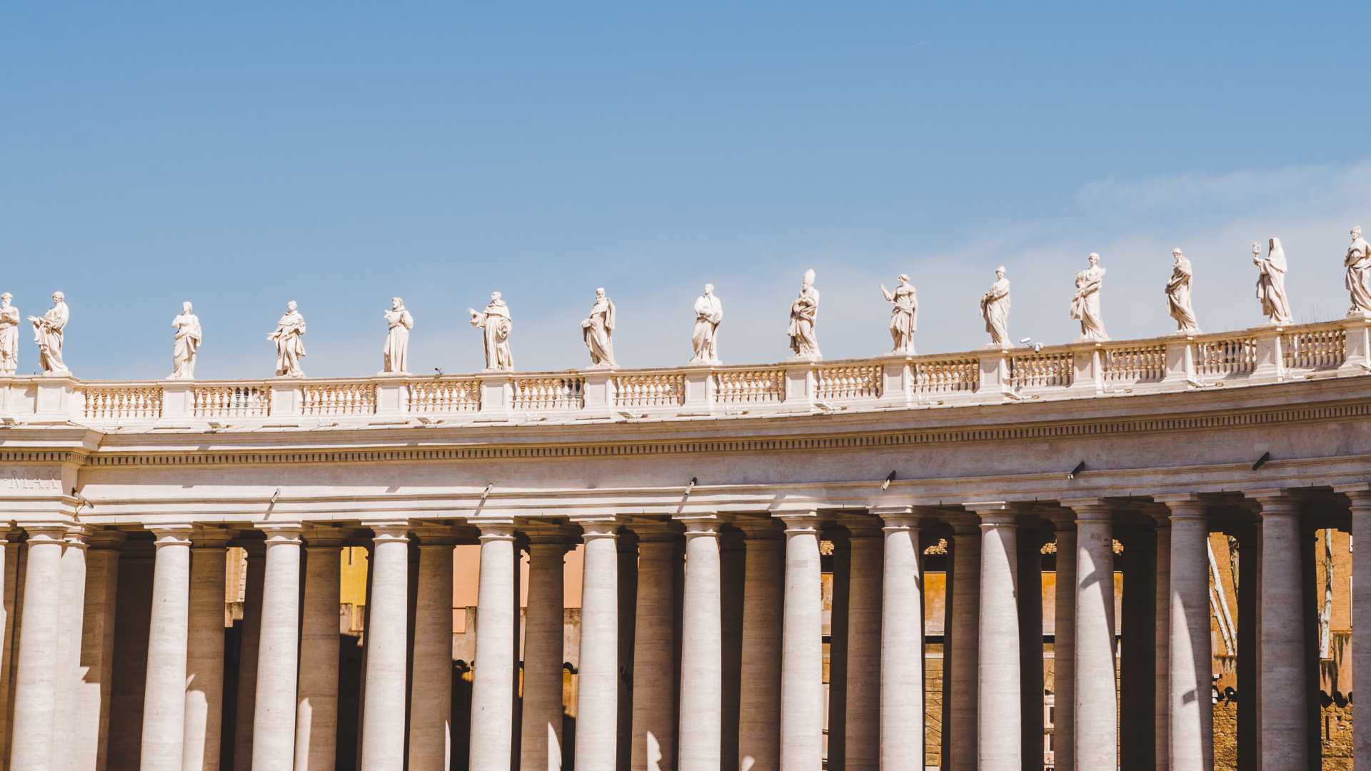 Standbeelden op grote zuilen onder een strakblauwe hemel op het Sint-Pietersplein, Vaticaanstad.